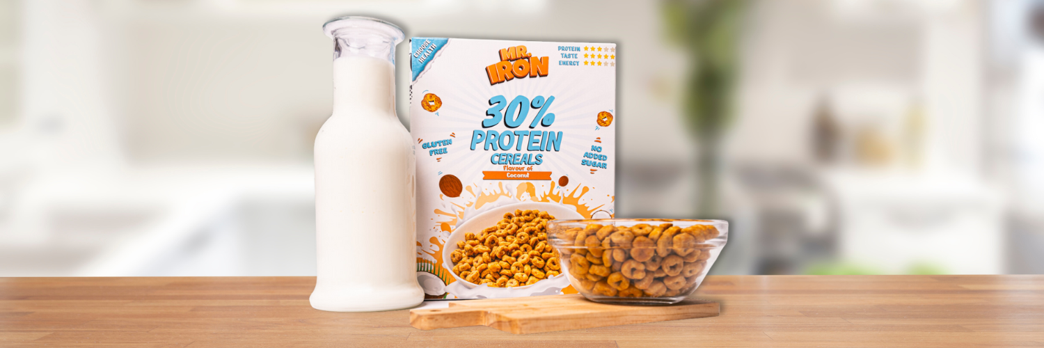 Cutie de cereale proteice Mr. Iron cu 30% proteine si aroma de cocos, fara gluten sau zahar adaugat, alaturi de o sticla de lapte si un bol plin, pe un blat de bucatarie.